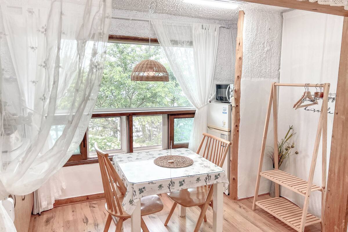 Una habitación acogedora con una mesa de comedor de madera, sillas, una pequeña cocina con refrigerador y un estante de madera.