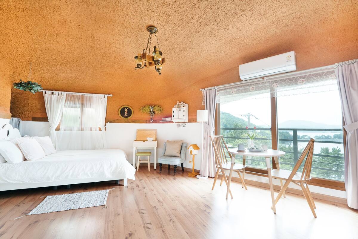 Un dormitorio con cama, escritorio, silla, mesa con lámpara y vistas al exterior.