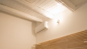 Una cama bien hecha con edredón marrón y manta blanca, cabecera de madera, mesita de noche con lámpara y unidad de aire acondicionado en la pared.
