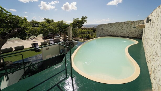 Una piscina con un muro de piedra y vista a un estacionamiento con un coche y mobiliario exterior.