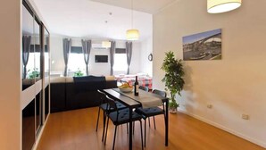 A modern living room with a dining area, a sofa, a table with chairs, a hanging light fixture, and a potted plant.