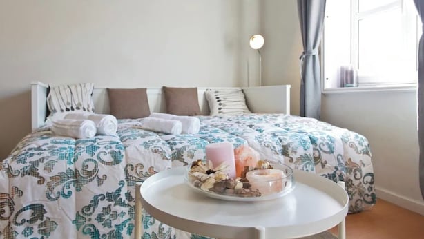 A neatly made bed with a patterned bedspread, a bedside table with a tray of snacks, and a bedside lamp.