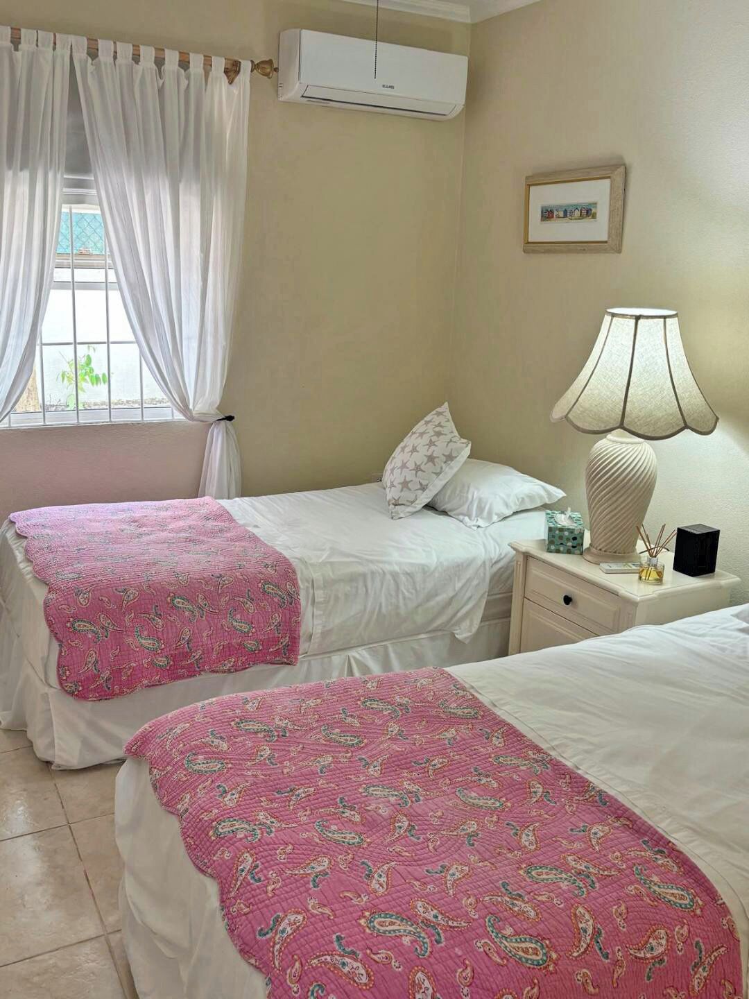 Ein Zimmer mit zwei Betten, jeweils mit einer rosa gemusterten Tagesdecke, einem Nachttisch mit Lampe und einer Schachtel sowie einer Klimaanlage an der Wand.