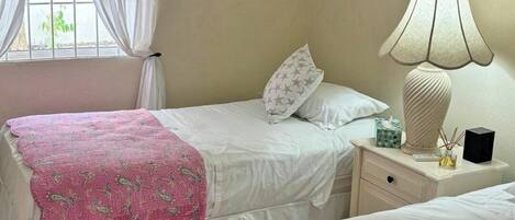 Ein Zimmer mit zwei Betten, jeweils mit einer rosa gemusterten Tagesdecke, einem Nachttisch mit Lampe und einer Schachtel sowie einer Klimaanlage an der Wand.