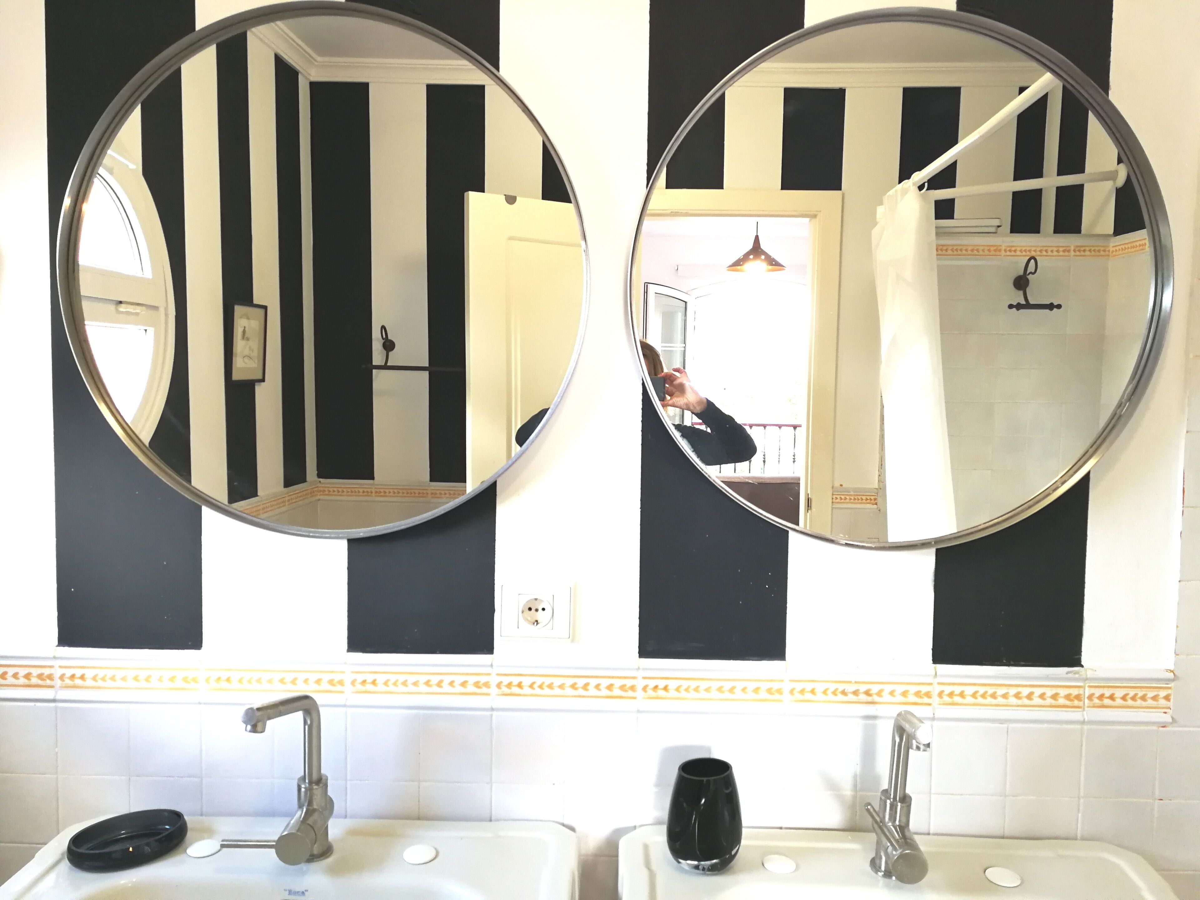 Dos espejos redondos colocados en una pared con azulejos a rayas negras y blancas, un lavamanos blanco con un jabonera negra, y un perchero con una toalla.