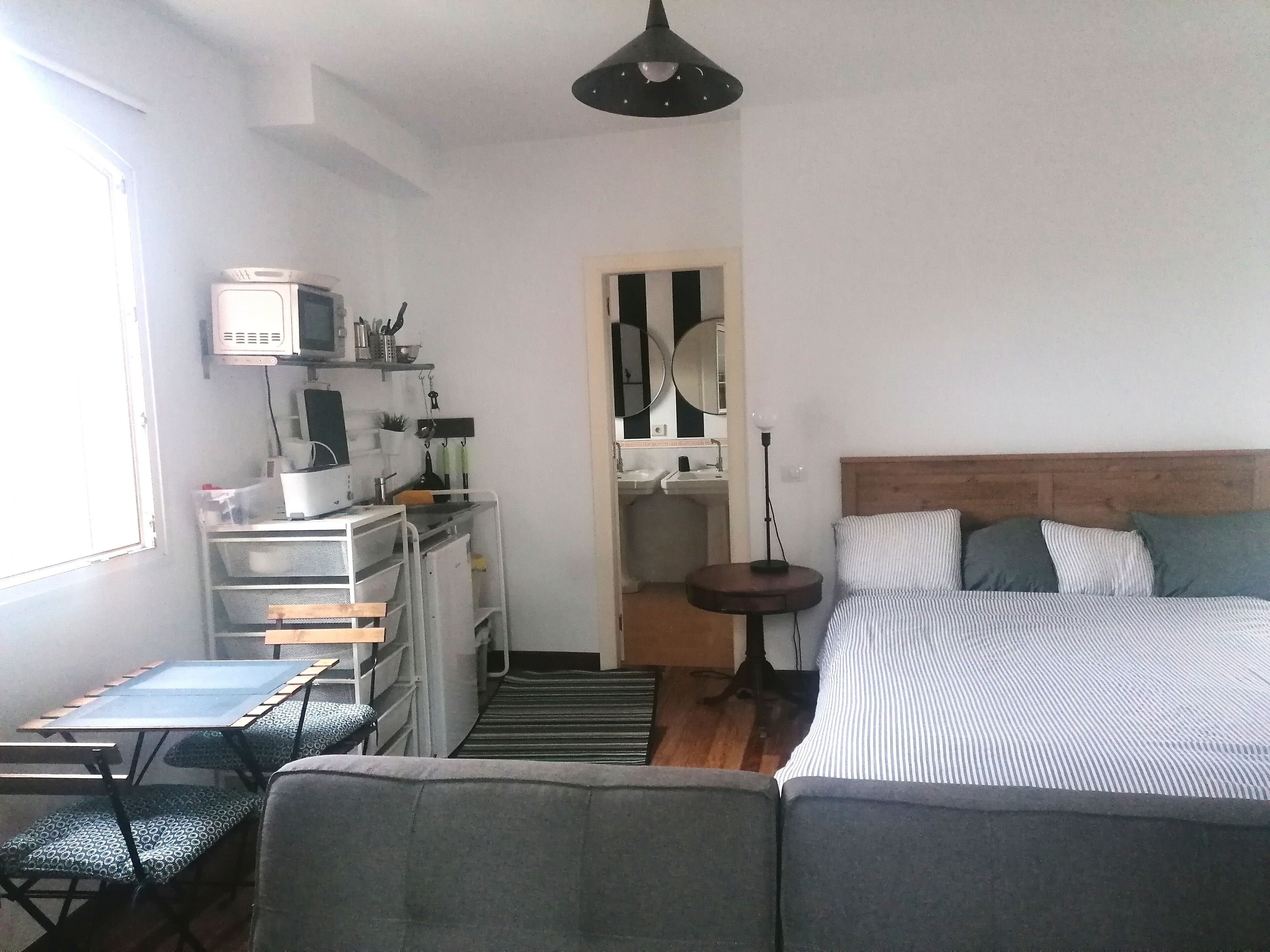 Un dormitorio compacto con cama, mesita de noche y una pequeña zona de escritorio con ordenador y espacio de almacenamiento.