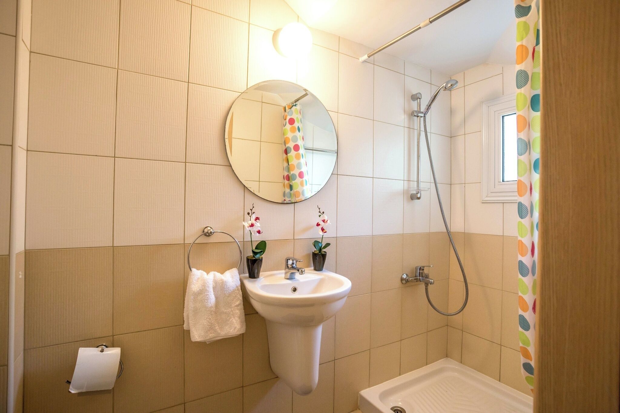 Et badeværelse med vask, spejl, håndklædetørre, toiletpapir, brusebad og et vindue.