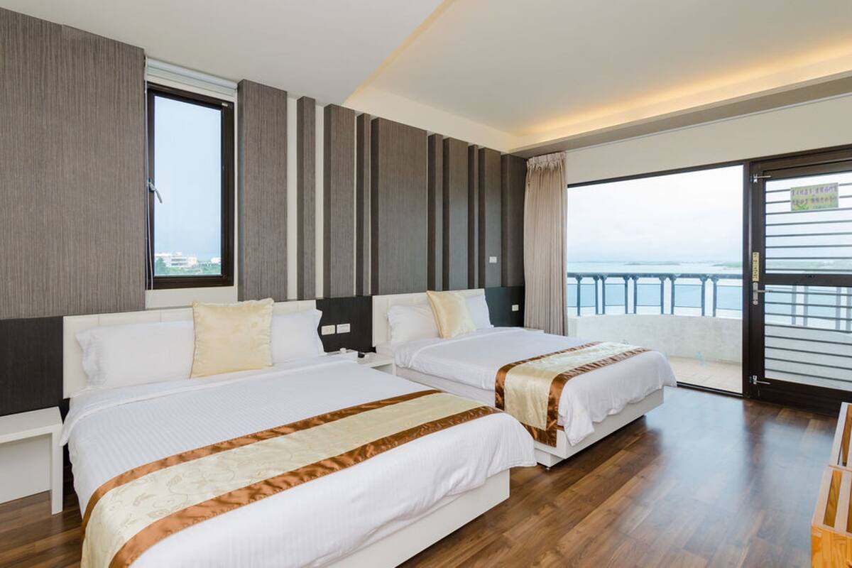 A hotel room with two beds, a large window, and a balcony with a view of the sea.