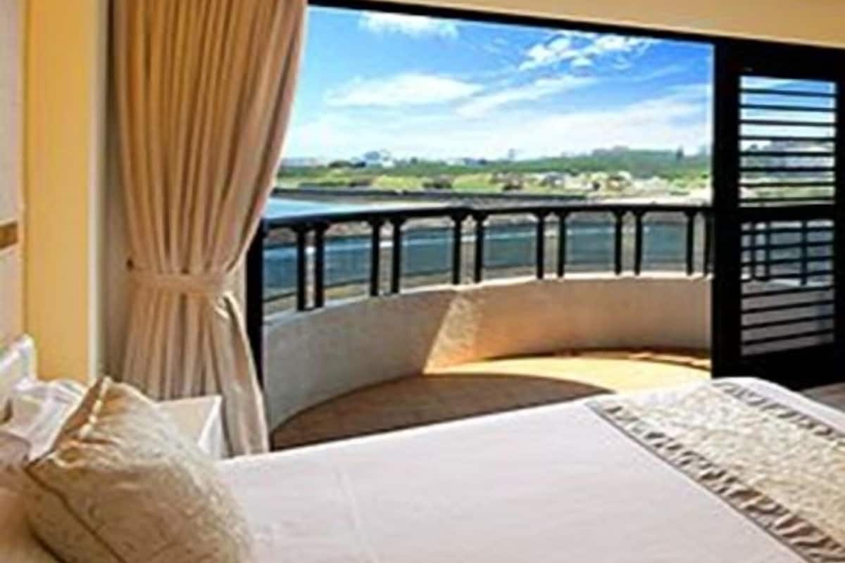 A bedroom with a large bed, a view of a body of water, and a balcony with a railing.
