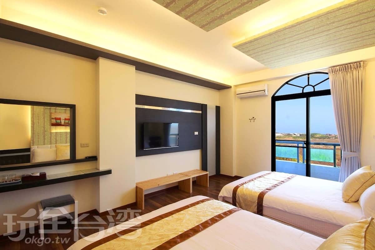 A hotel room with a large window, a flat-screen TV, a bench, and two beds.