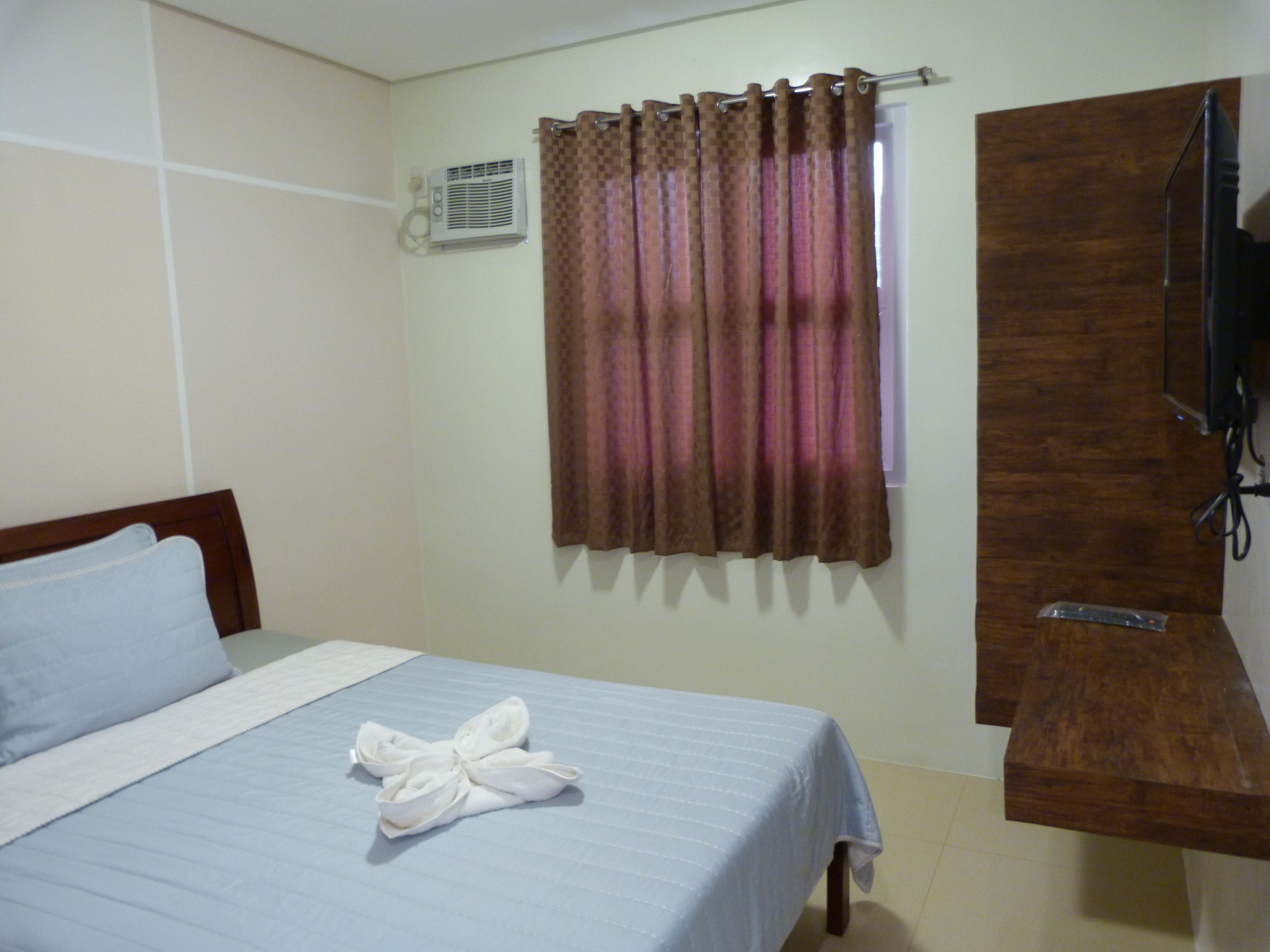 A hotel room with a bed, a TV mounted on a wooden panel, and a window with curtains.