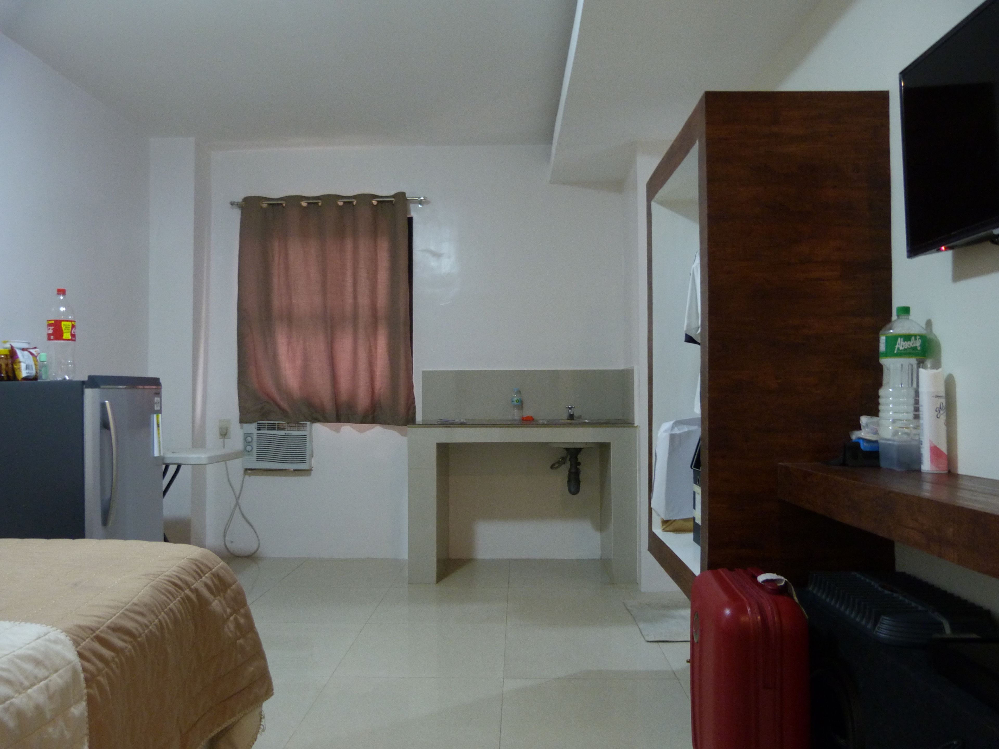 A hotel room with a bed, a desk with a mirror, a sink, a television, and a red suitcase.