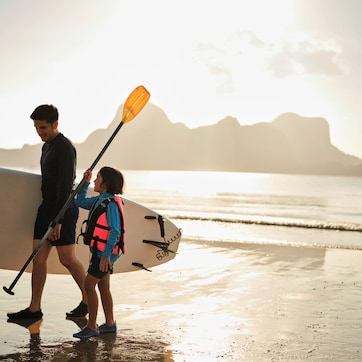 A person and a child walking on a beach with a surfboard and paddleboard.