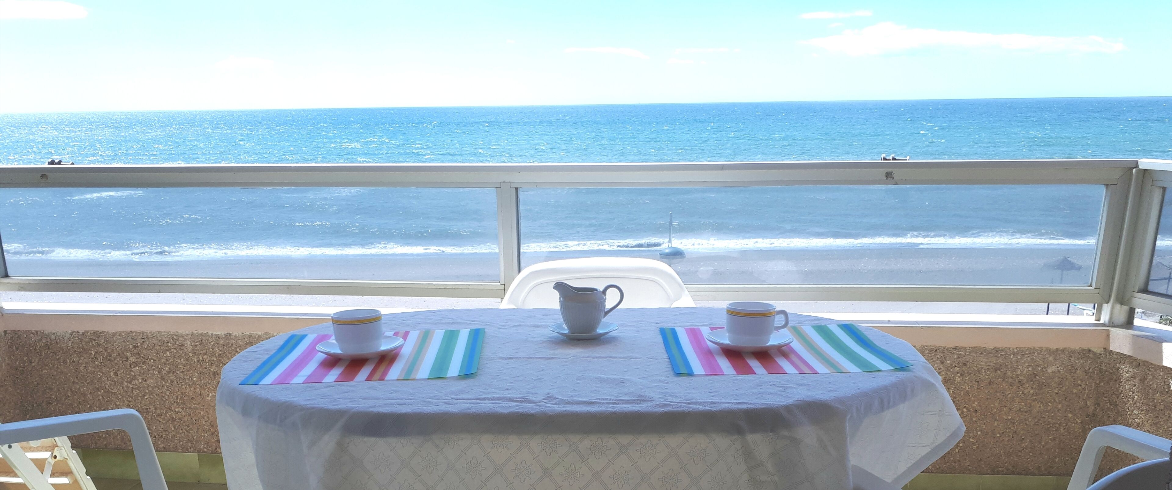 A table with a white tablecloth and two white cups on striped placemats, overlooking a beach and ocean.
