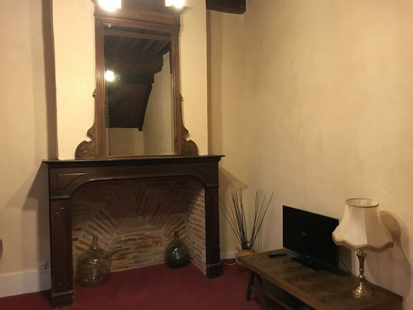 A room with a brick fireplace, a wooden mantel, a mirror above it, a television on a wooden stand, and a lamp on a side table. - Hotel Raymond VII (Cordes-sur-Ciel)