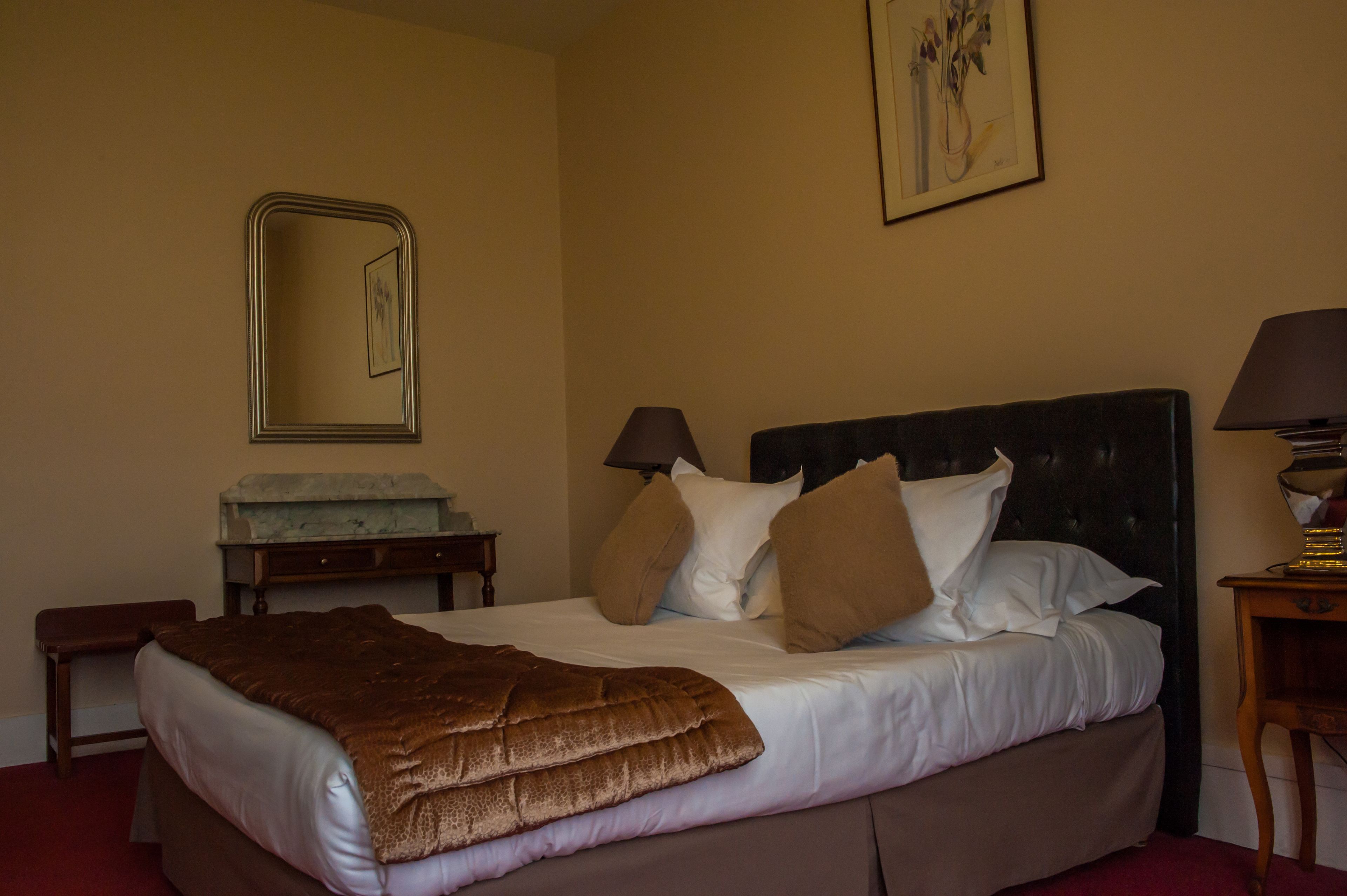 A neatly made bed with a brown blanket, two bedside lamps, a nightstand with a lamp, a small bench, and a framed picture on the wall.