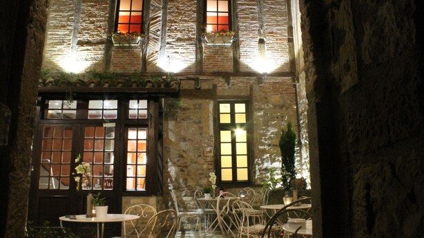 Un área de café al aire libre con paredes de piedra, varias mesas y sillas, y plantas en macetas.