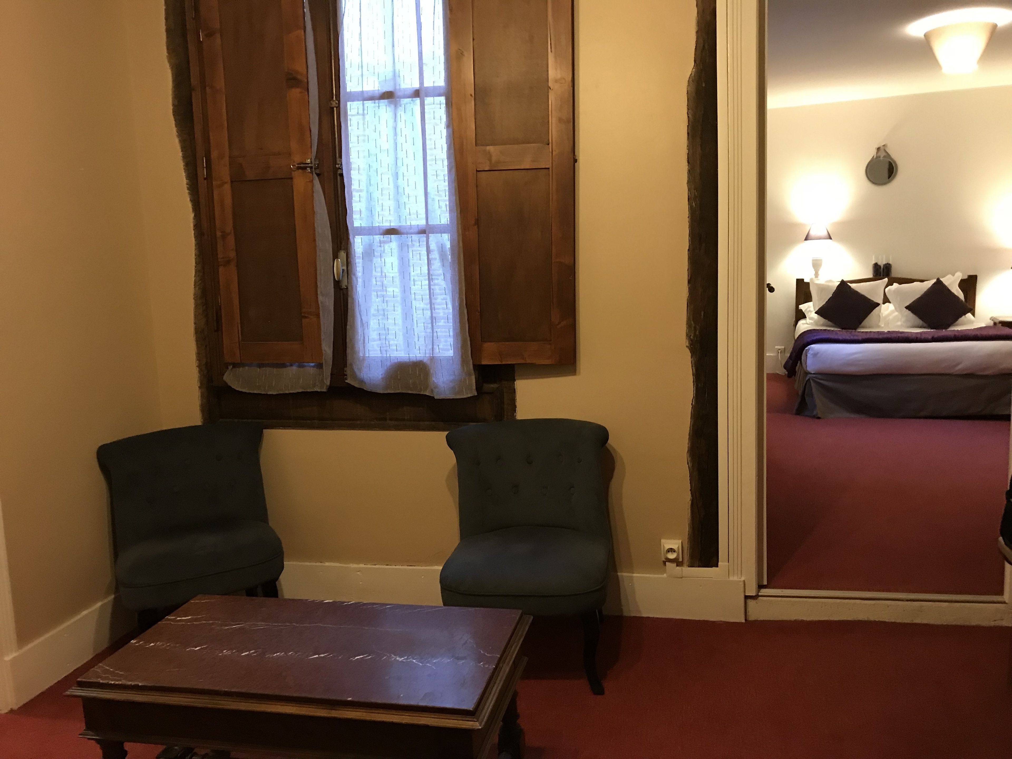 A room with two green armchairs, a wooden table, a window with wooden shutters, and a bed with a purple blanket and pillows.