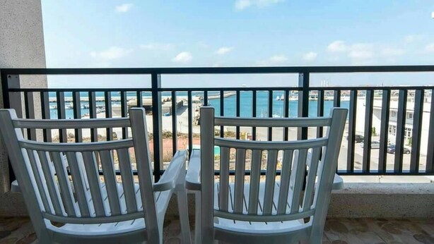 A balcony with two white chairs and a table, overlooking a coastal area with buildings and a body of water. - Longman Villa (Huxi)