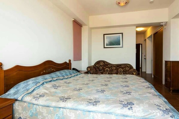 Un dormitorio con una cama grande, un cabecero de madera, una colcha estampada con motivos florales, un sofá marrón, un cuadro enmarcado en la pared y un mueble de madera.