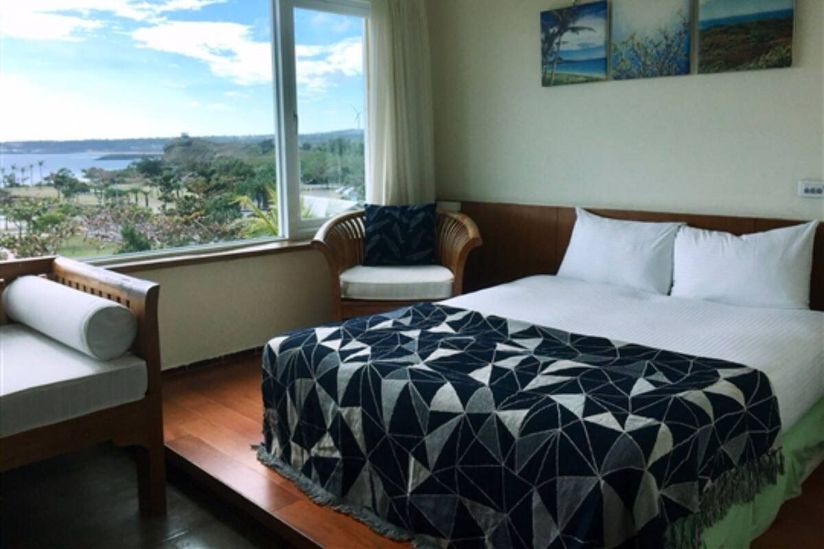 A hotel room with a bed, a chair, a window with a view of the landscape, and artwork on the wall.
