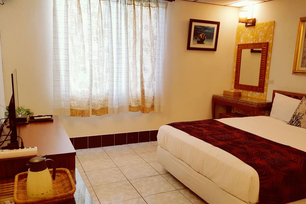A hotel room with a bed, a desk, a chair, a lamp, a plant, and a window with curtains.