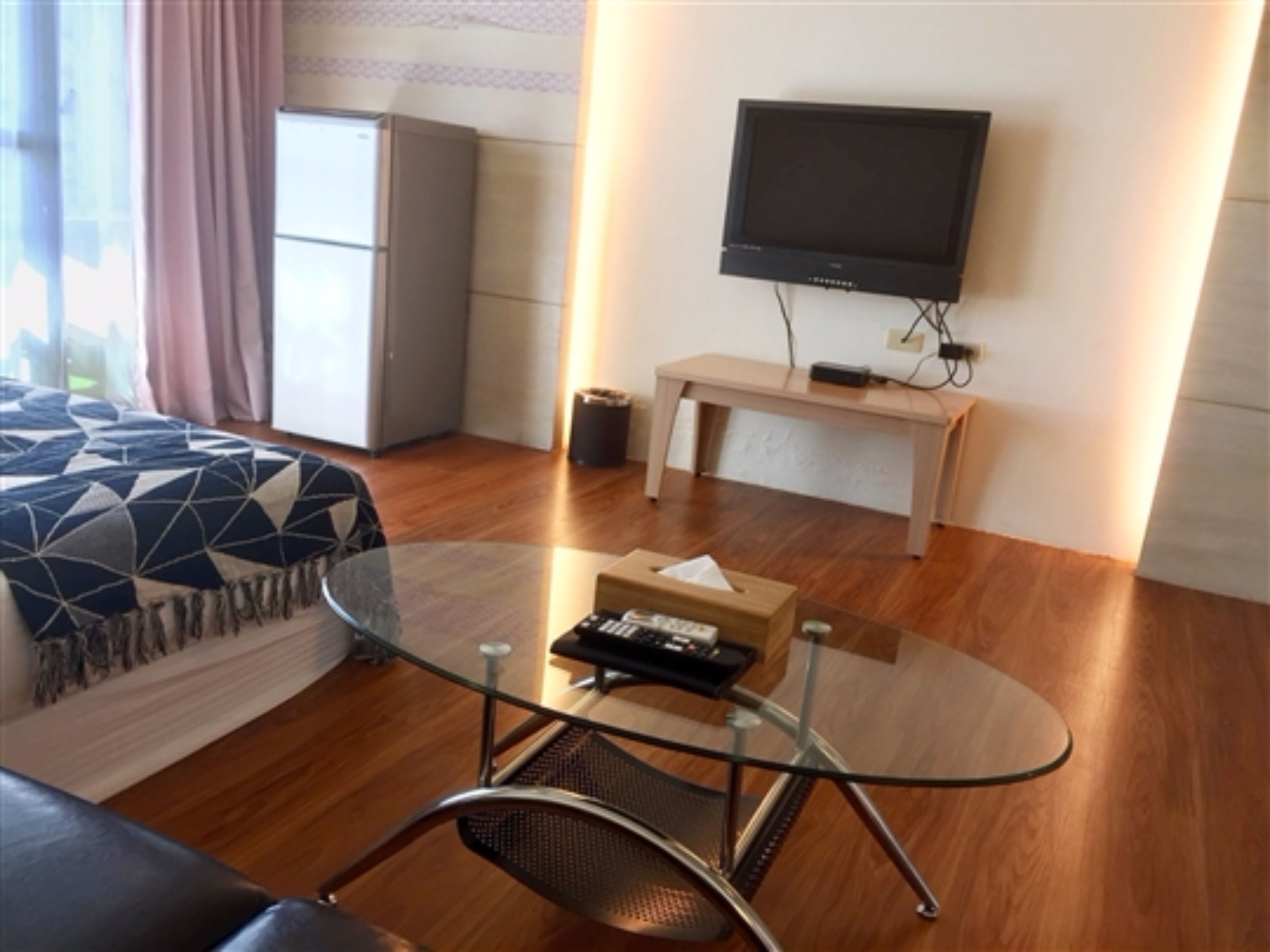 A modern hotel room with a glass coffee table, a sofa, a TV mounted on the wall, and a wooden floor.