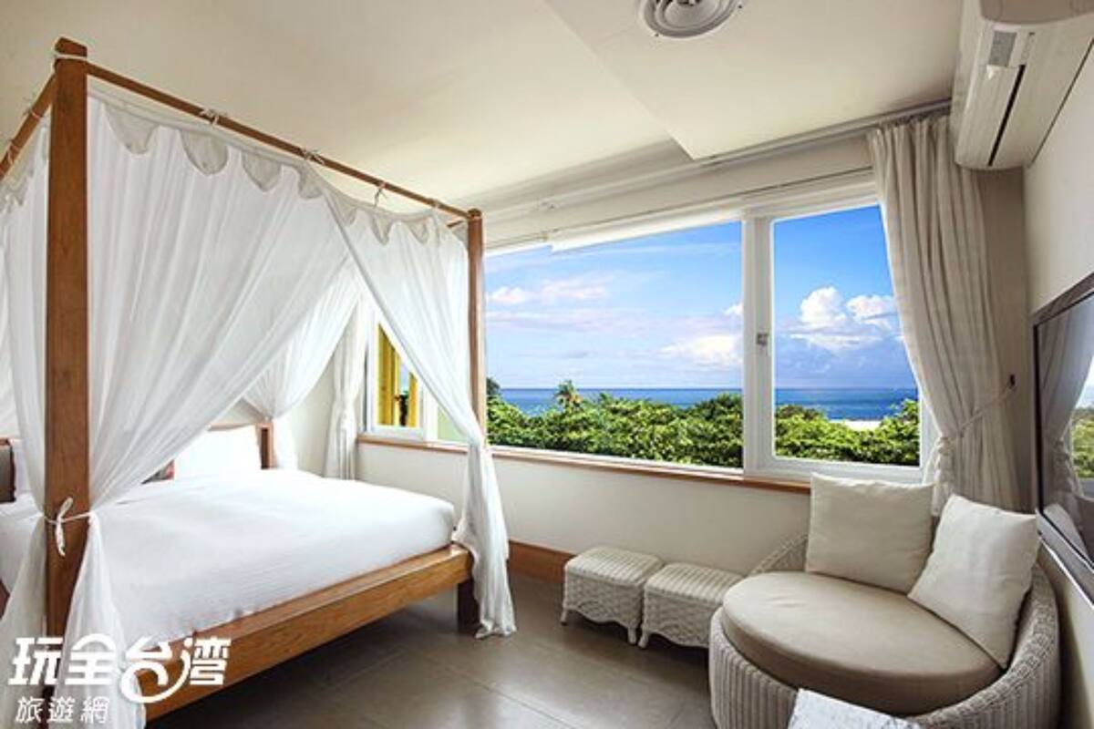 A bedroom with a four-poster bed, a small seating area, and a large window with a view of the ocean.