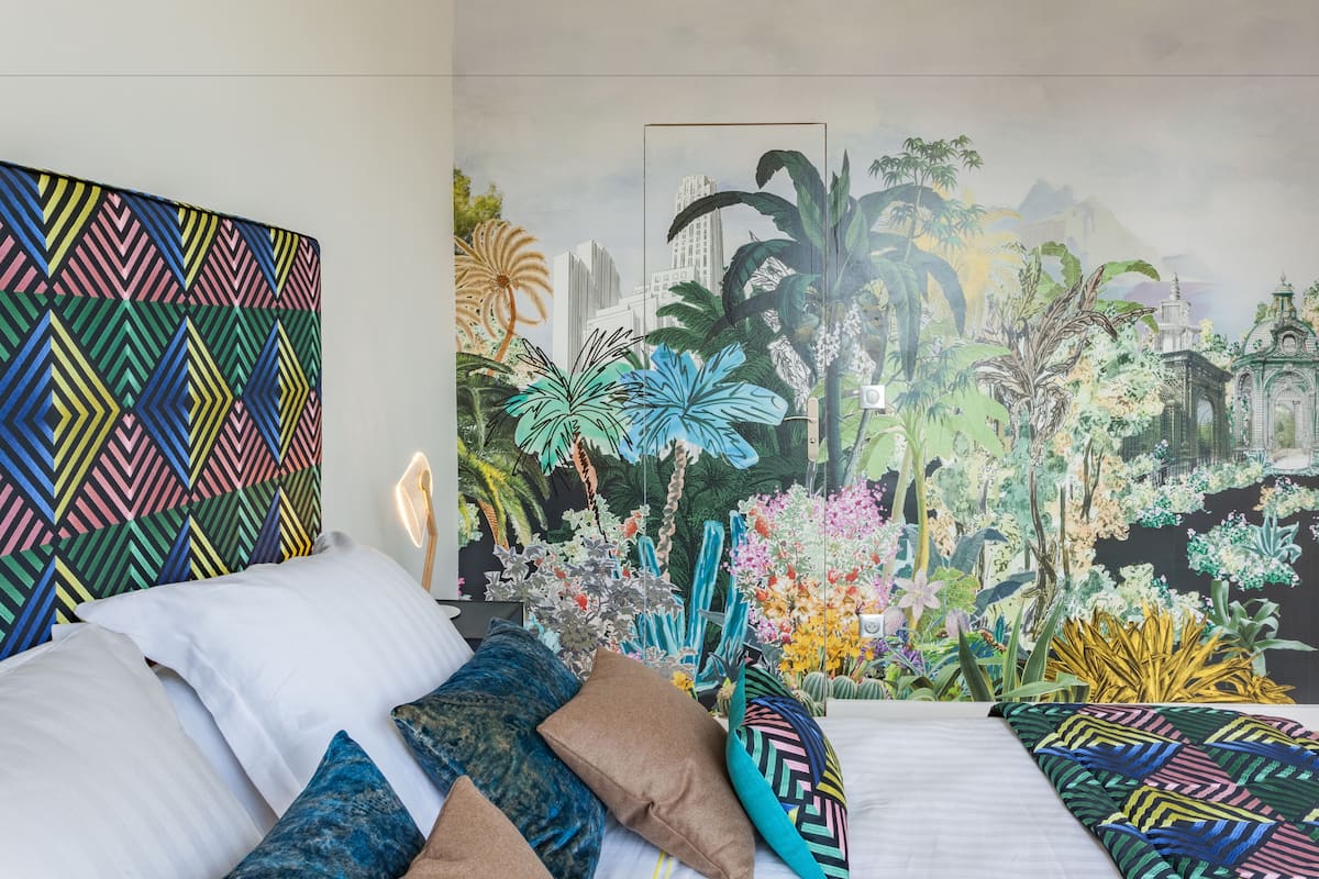 Un dormitorio con cama, almohadas y un gran mural de un paisaje urbano y plantas tropicales.