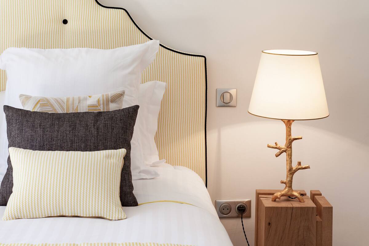Una cama bien tendida con ropa de cama a rayas, una mesita de noche de madera con una lámpara e interruptor de luz en la pared.