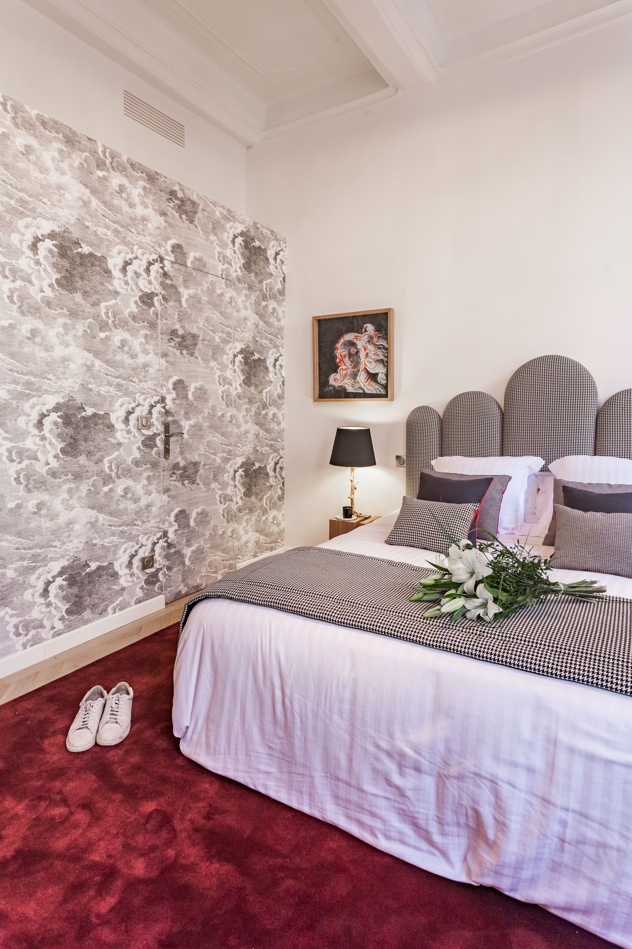 A bedroom with a stone wall, a bed with a white and black checkered bedspread, a nightstand with a lamp, and a pair of shoes on the floor.