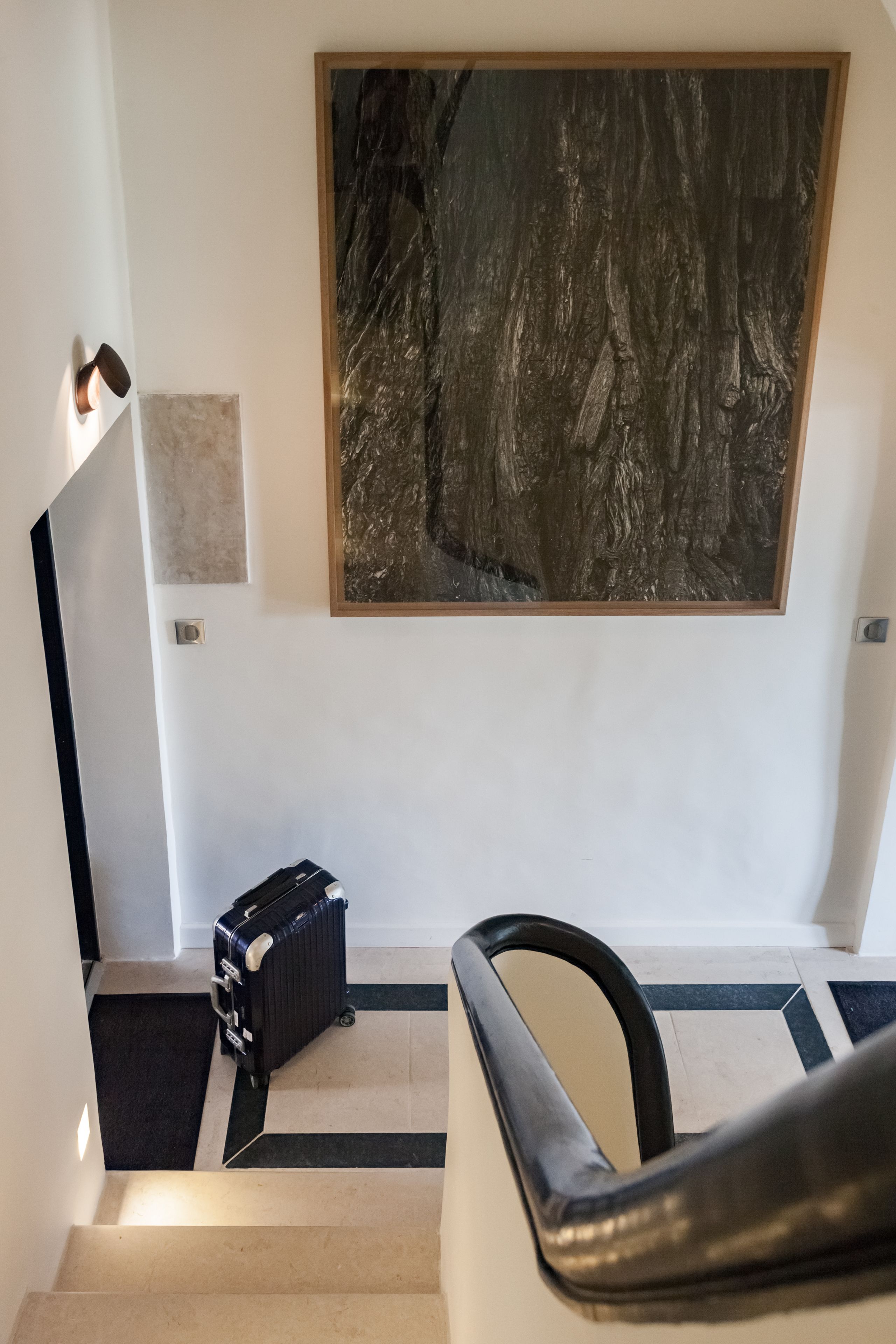Eine Treppe mit einem schwarzen Koffer auf der Zwischenebene, einem gerahmten Kunstwerk darüber und einer Wandleuchte.