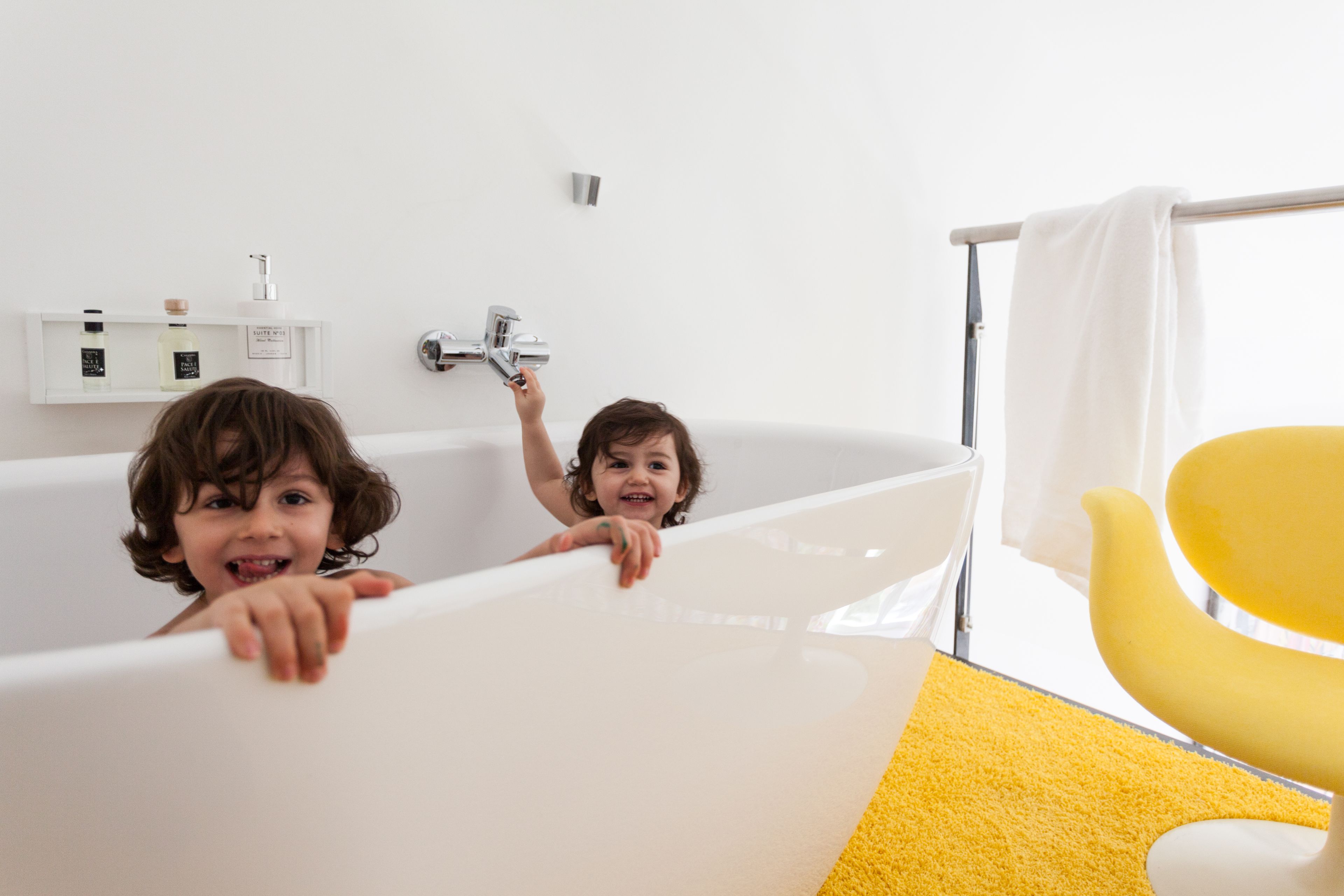 A bathtub with two children in it, a yellow chair, and a white towel hanging on a rack.