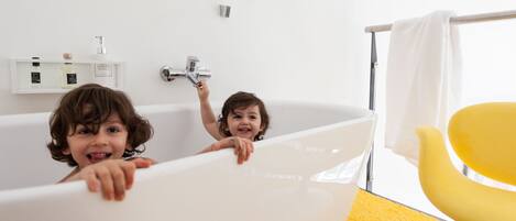 A bathtub with two children in it, a yellow chair, and a white towel hanging on a rack.