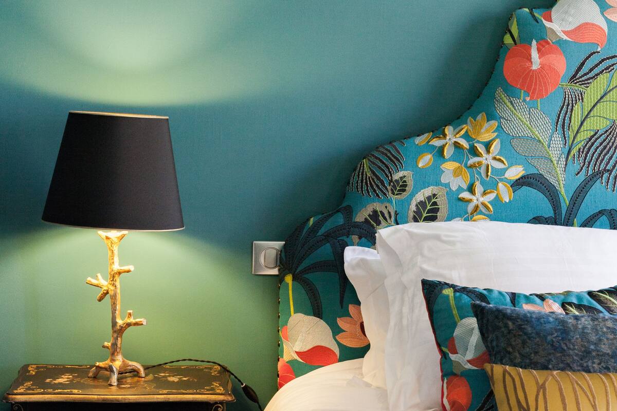 Una cama con colcha estampada, una lámpara negra sobre una mesita de noche y una pared de color verde azulado.