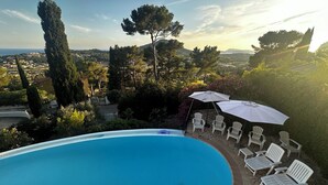 Ein Swimmingpool mit weißen Liegestühlen und Sonnenschirmen, umgeben von Bäumen und Blick auf eine Stadt und Berge.