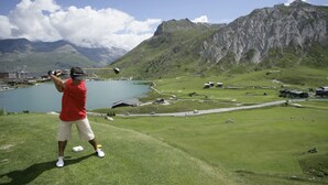 Un uomo gioca a golf su un pendio erboso, con un lago e montagne sullo sfondo.