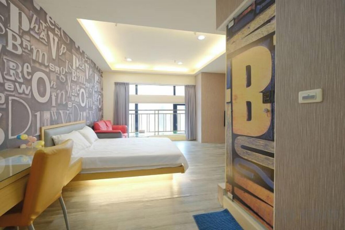 A hotel room with a large bed, a desk, a chair, and a view of the city through the window.