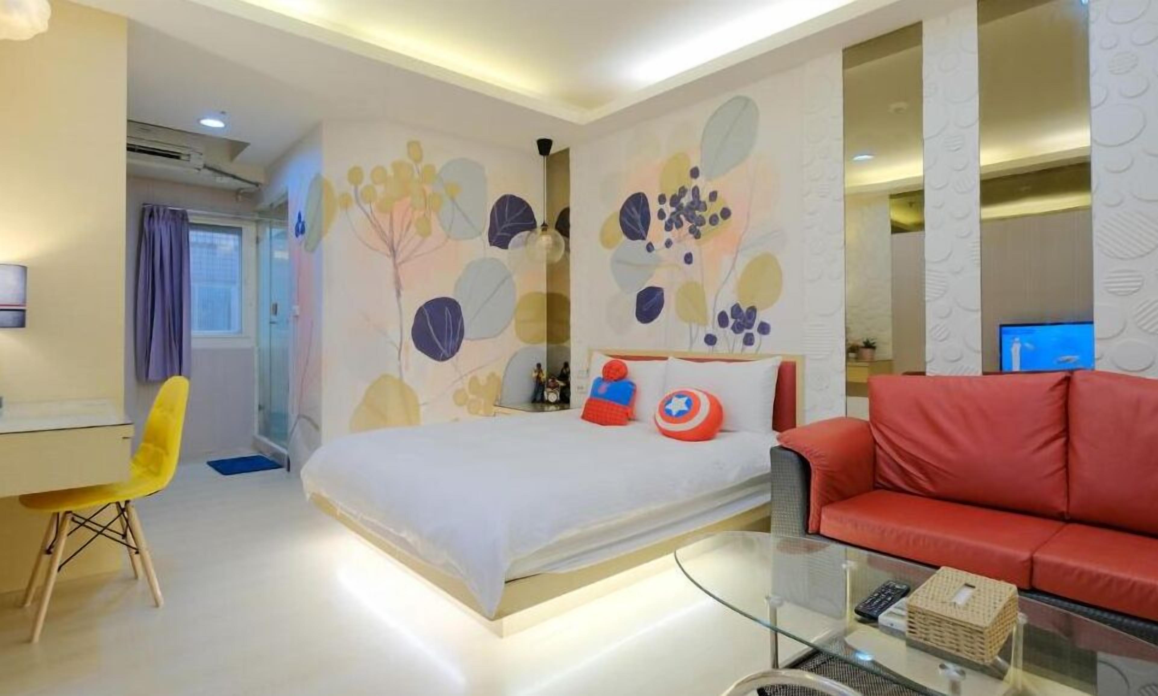 A modern hotel room with a bed, a red sofa, a glass coffee table, and a desk with a chair.