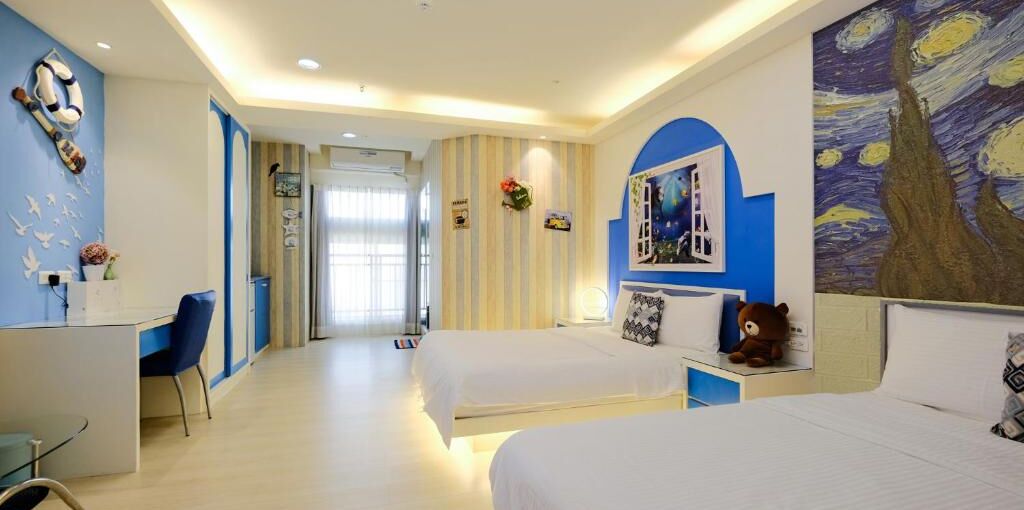 A hotel room with two beds, a desk, and a chair. There is a painting on the wall and a teddy bear on the bed.