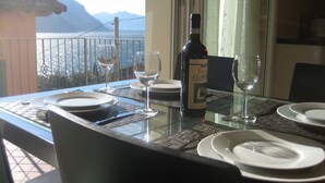 A dining table with plates, glasses, and a bottle of wine, set on a balcony with a view of mountains and the sea.