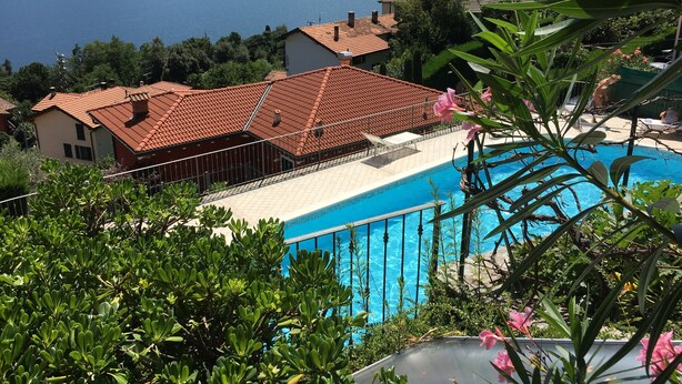 A pool surrounded by lush greenery, a red-tiled house, and a view of a lake with mountains in the background.