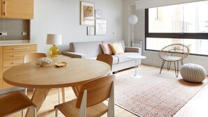 Una sala de estar moderna con una mesa redonda de madera, un sofá y una silla. Hay un ventanal grande que proporciona luz natural y vistas al exterior.