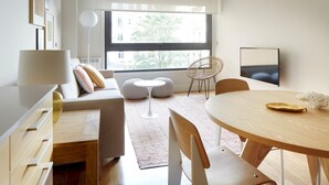Una sala de estar moderna con un sofá, una mesa de comedor redonda, un televisor y un ventanal con vistas a edificios y árboles.
