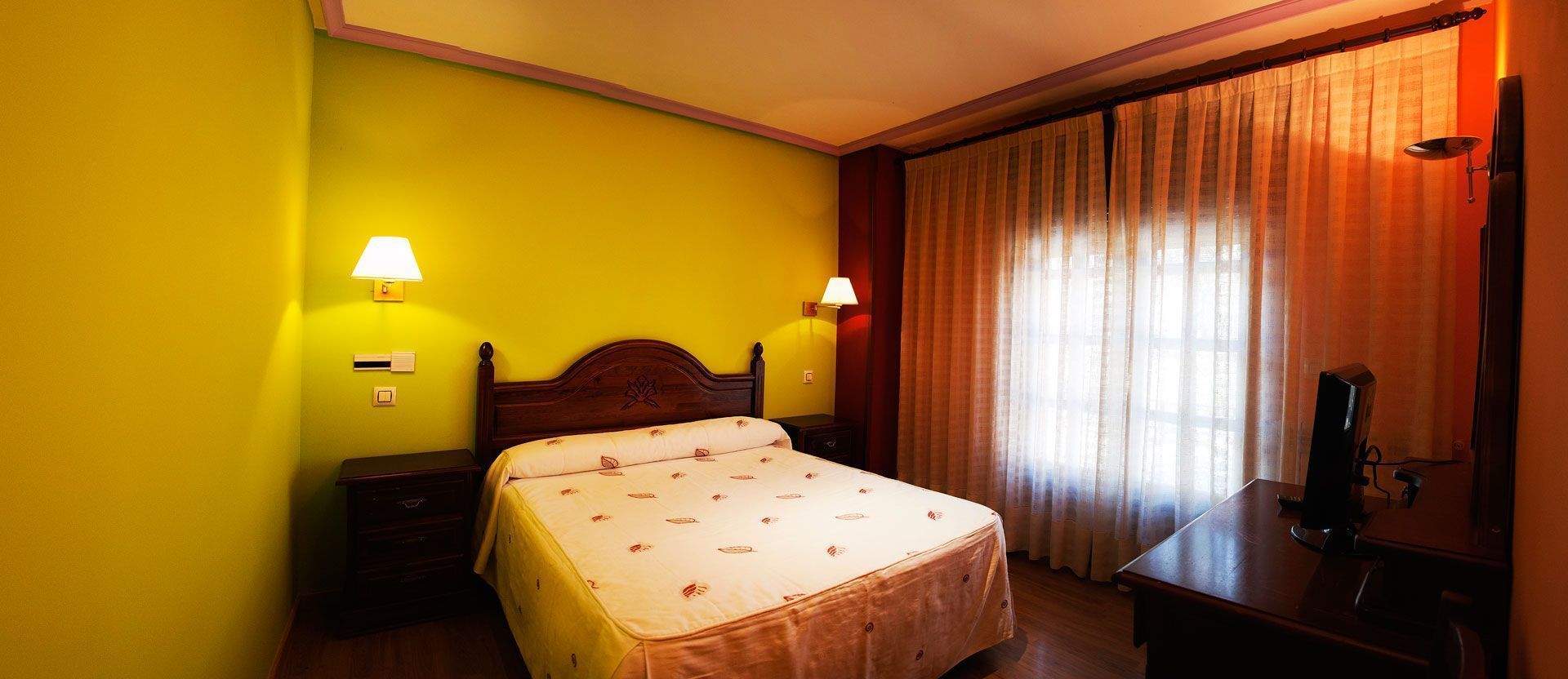 Habitación de hotel con cama, dos lámparas de noche, un escritorio con computadora y ventana con cortinas.