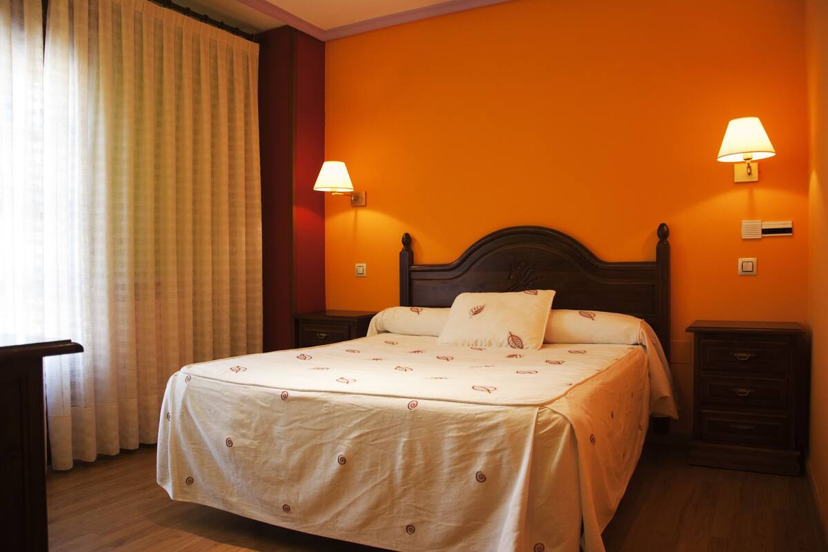 Una habitación de hotel con una cama grande, dos mesitas de noche, una lámpara y un ventanal con cortinas.