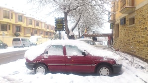 A red car covered in snow parked on a street with buildings in the background. - Hotel Rural Tierra de Lobos (Puebla de Sanabria)