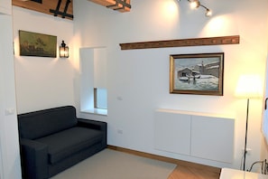 A modern living room with a dark sofa, a small window, a radiator, and a painting of a snowy scene.