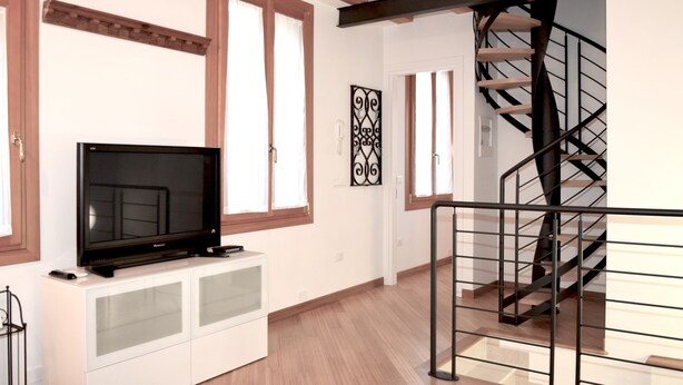 A modern living room with a flat-screen TV on a white cabinet, wooden floors, a staircase with a metal railing, and decorative wall art.