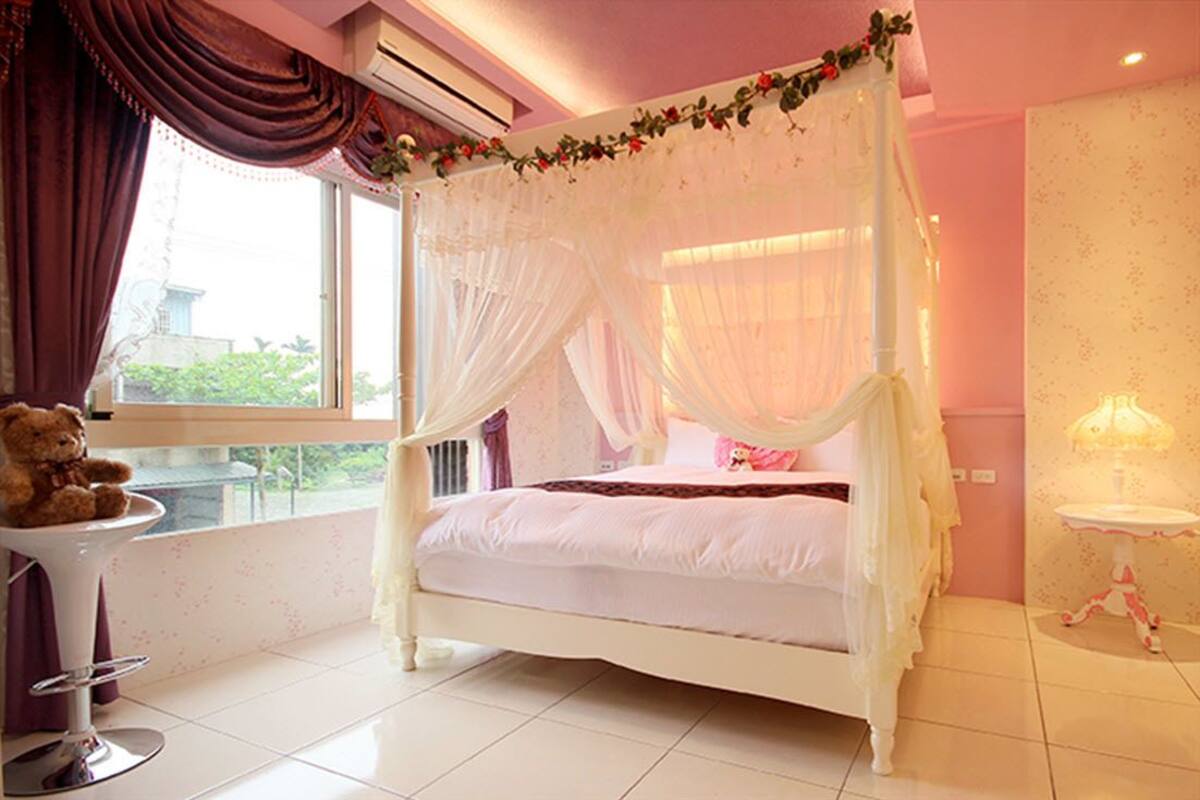 A four-poster bed with a canopy, a bedside table with a lamp, and a teddy bear on a chair.